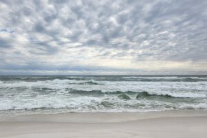 Overcast sky above a gray-blue ocean with white-capped waves rolling onto a pale sandy shore. For the post Strengths, Personal and Professional